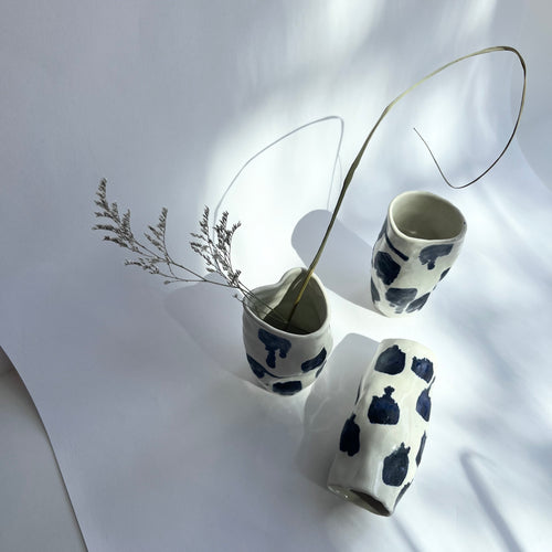 Three ceramic vases, each appearing to be slightly squished, sit on a piece of white paper. One is toppled over and one has dried flowers in it. Each one is white with navy splotches.