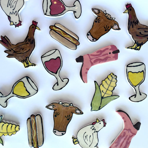 Various painted ceramic magnets are place on a white backdrop. Chickens in white and brown, wine glasses in red or white, a cute brown cow, corn, hot dogs, and pink cowboy boots!